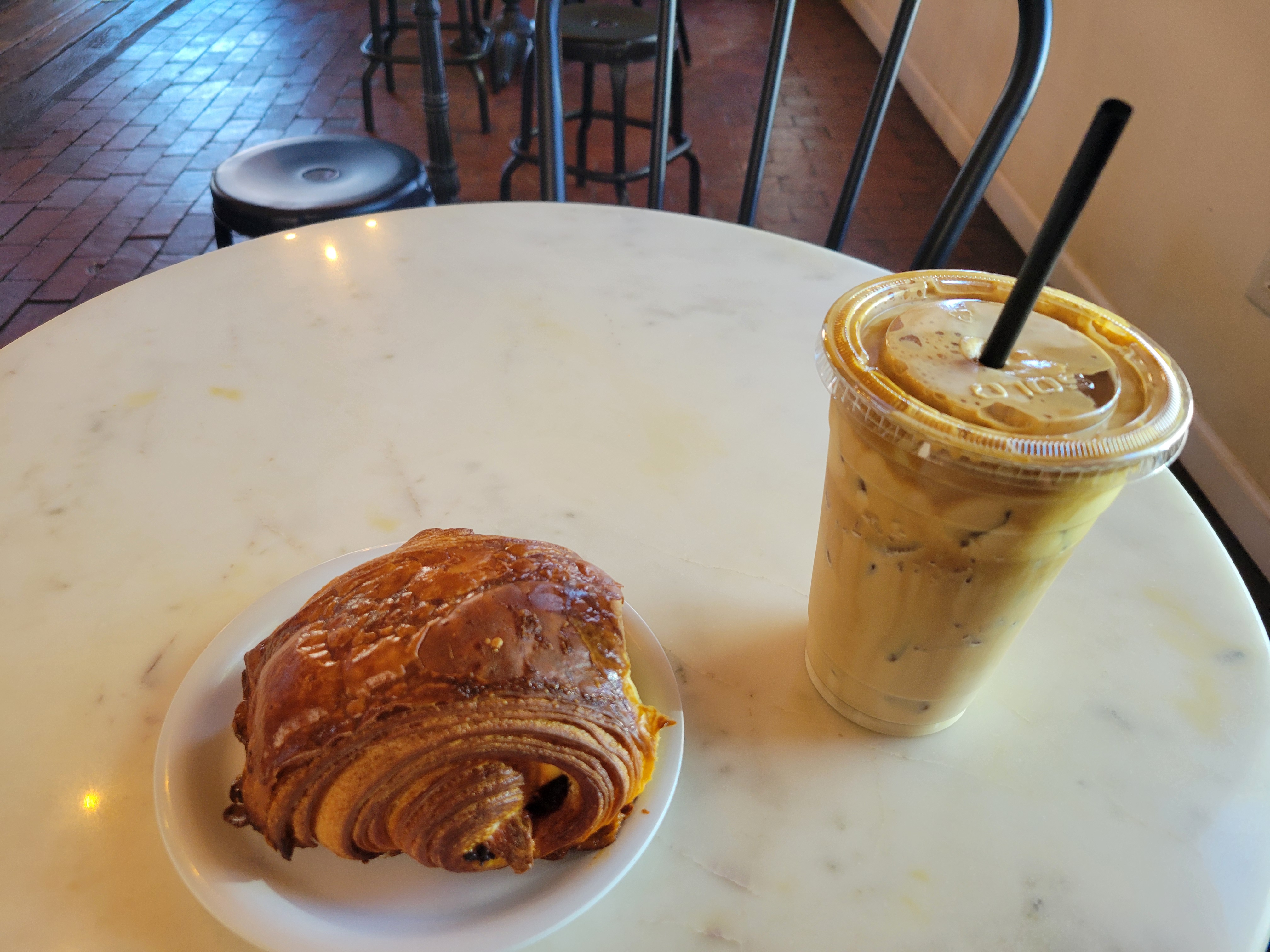Plastic cup of coffee and a croissant on a plate on a table.